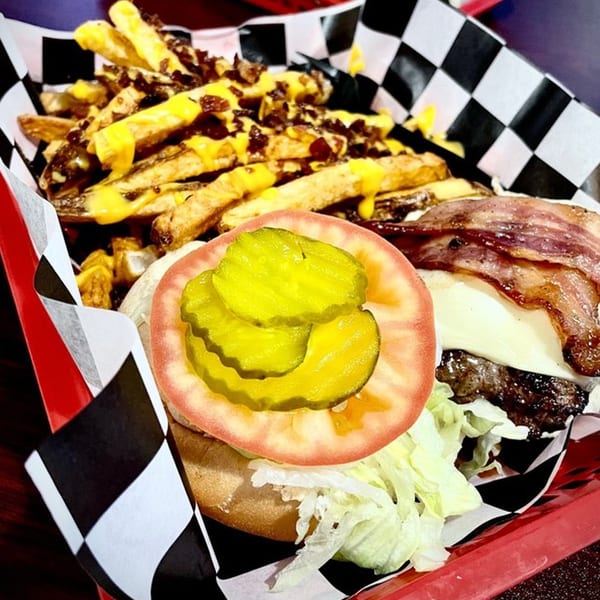 Bacon cheeseburger with lettuce, tomato, and pickles, served with cheesy bacon fries in a red basket.